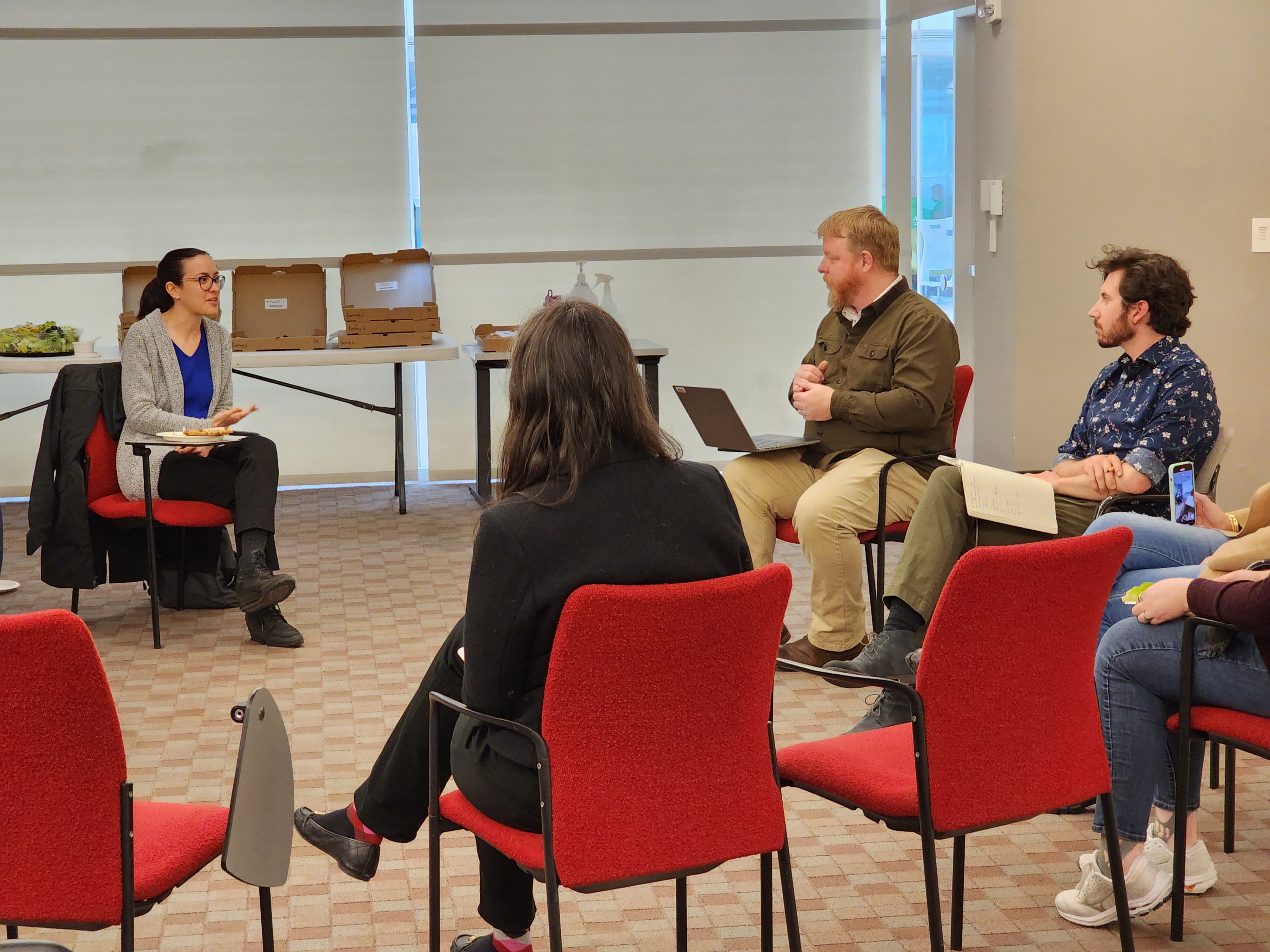 A woman sitting in a chair is in conversation with a man in a nearby chair. They are both looking at eachother. A few other people are seated nearby.