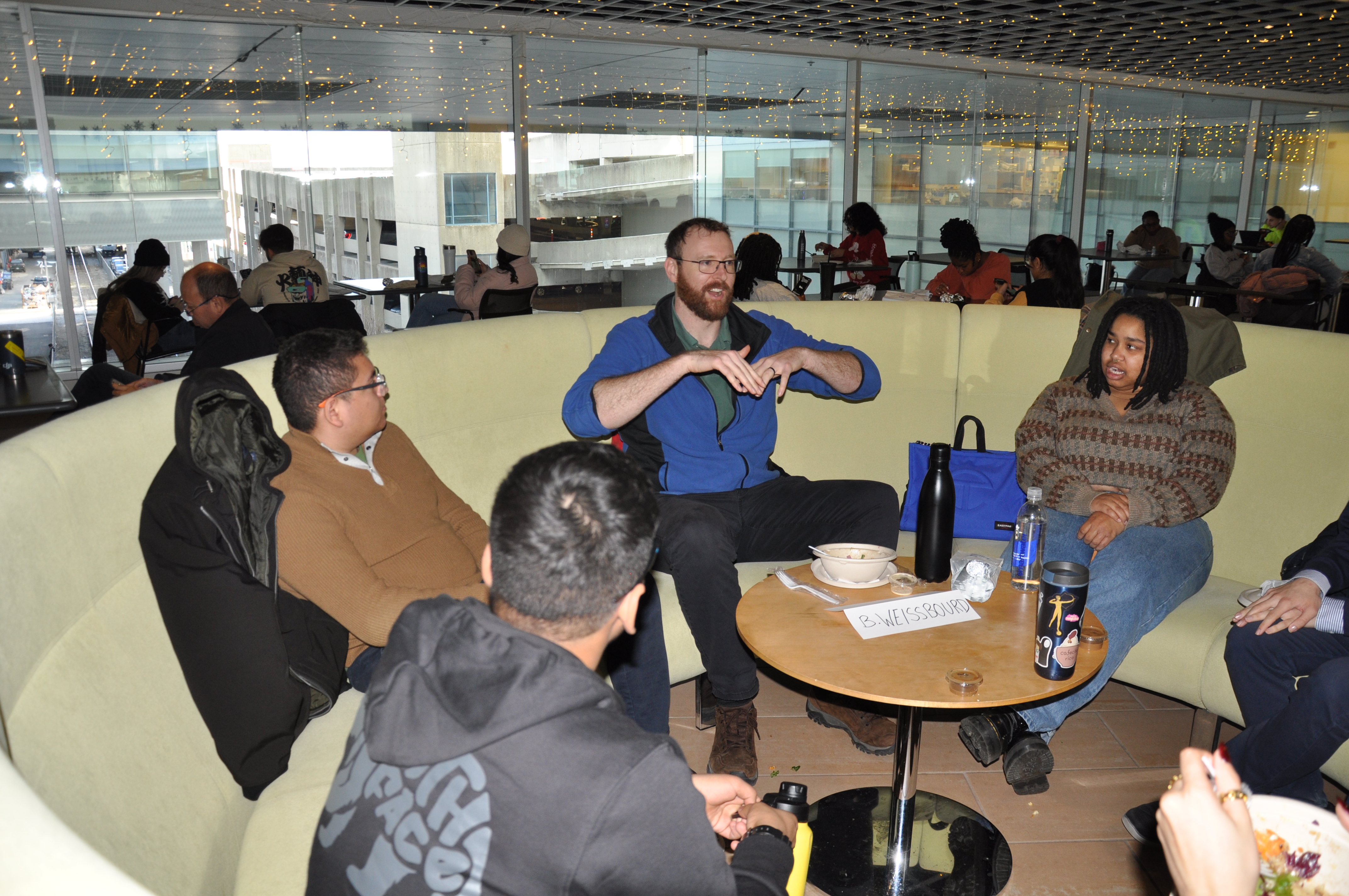 Brady Weissbourd sits on a large, circular yellow couch in MIT Building 46 with QMW students on the couch next to him on both sides.