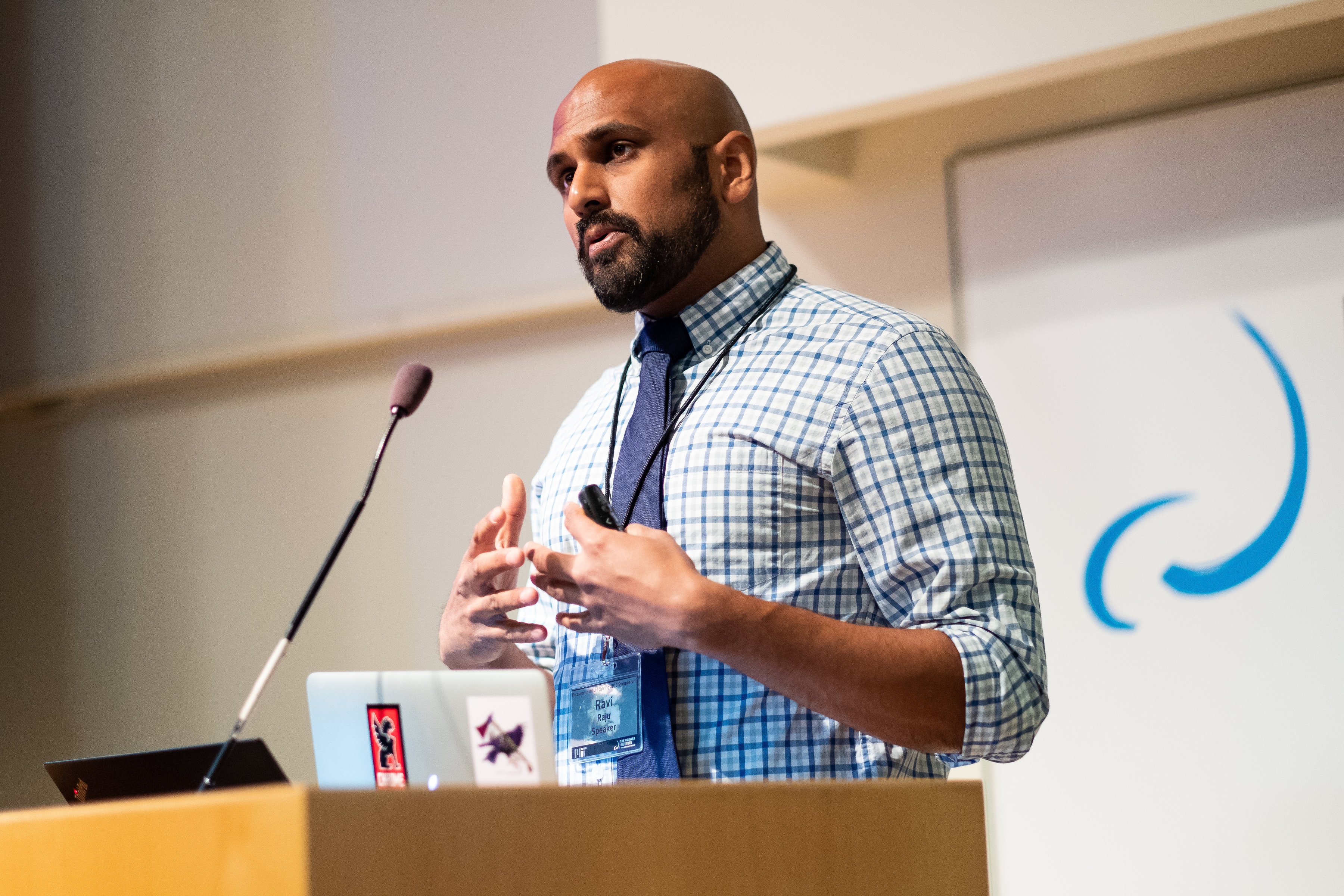 Ravi Raju stands at a podium. A blue and white Picower Institute logo is behind him as a backdrop.