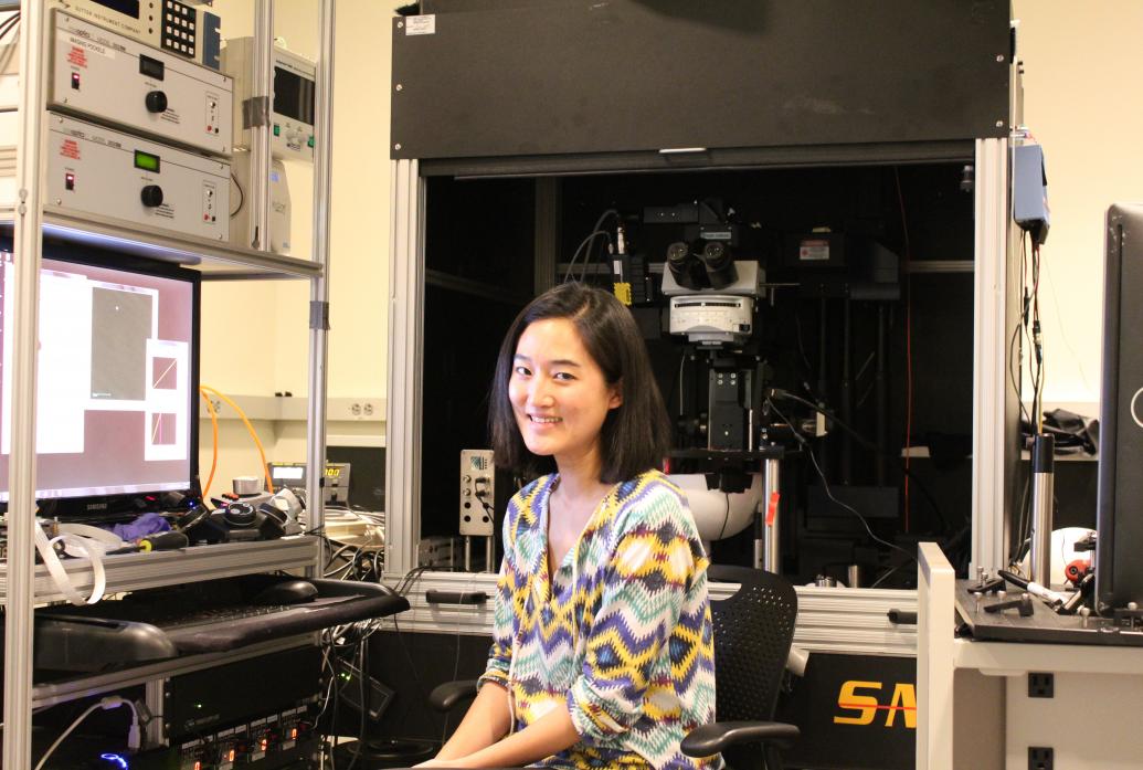 A woman sits at a lab computer with a microscope in the background