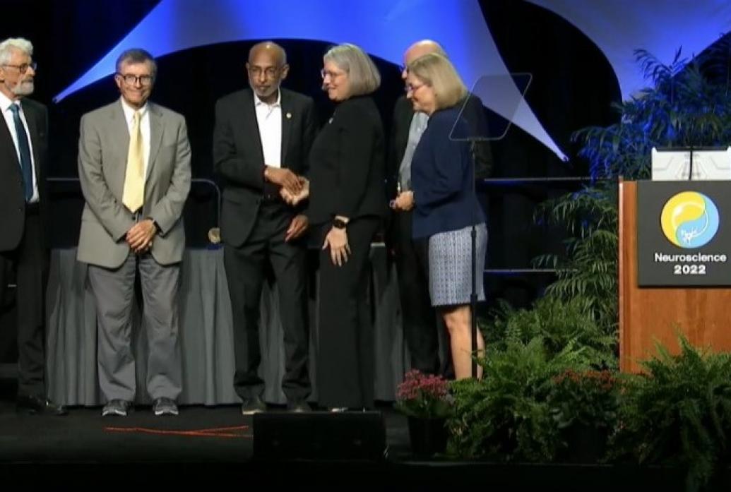 On a stage with plants and a blue background with a podium labeled Neuroscience 2022, Emery Brown receives a handshake from a woman after accepting a neuroscience prize