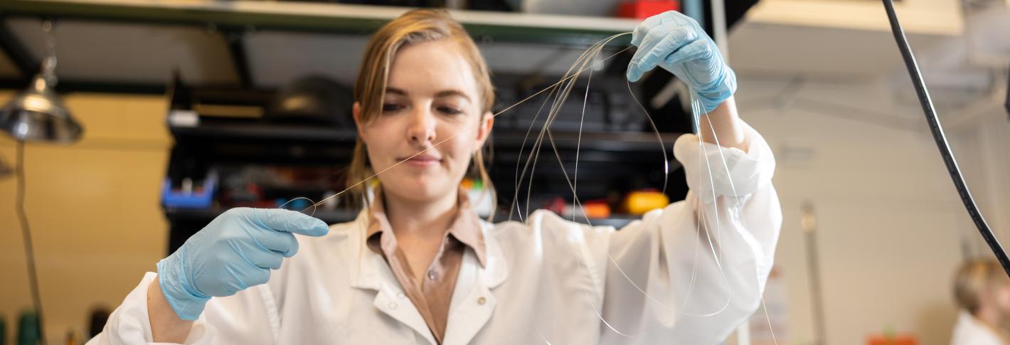 Indie garwood, wearing latex gloves and a lab coat, streches out a fiber in the lab.