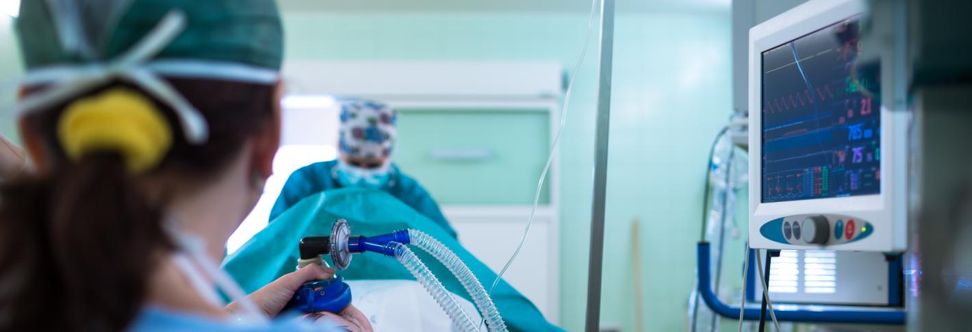 An operating room scene shows a patient on a table. Our perspective is from behind the anesthesiologist who holds a mask on the patient's face and watches a monitor with a bunch of indicators. A surgeon stands out of focus on the far end of the patient.