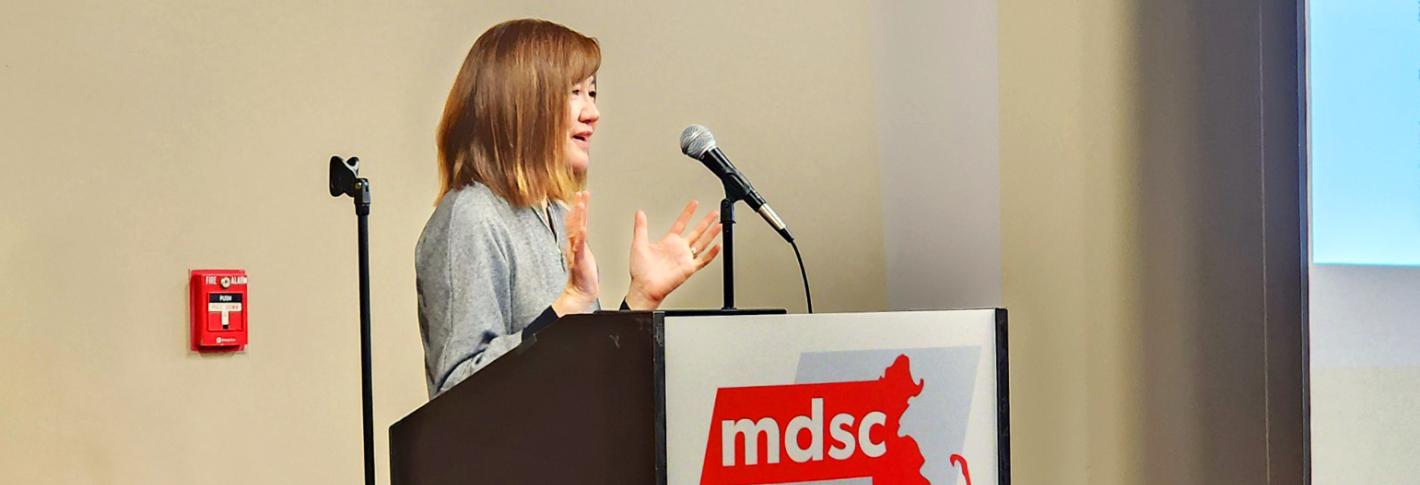 Li-Huei Tsai smiles and gestures as she stands at a podium with a placard displaying the Massachusetts Down Syndrome Congress logo