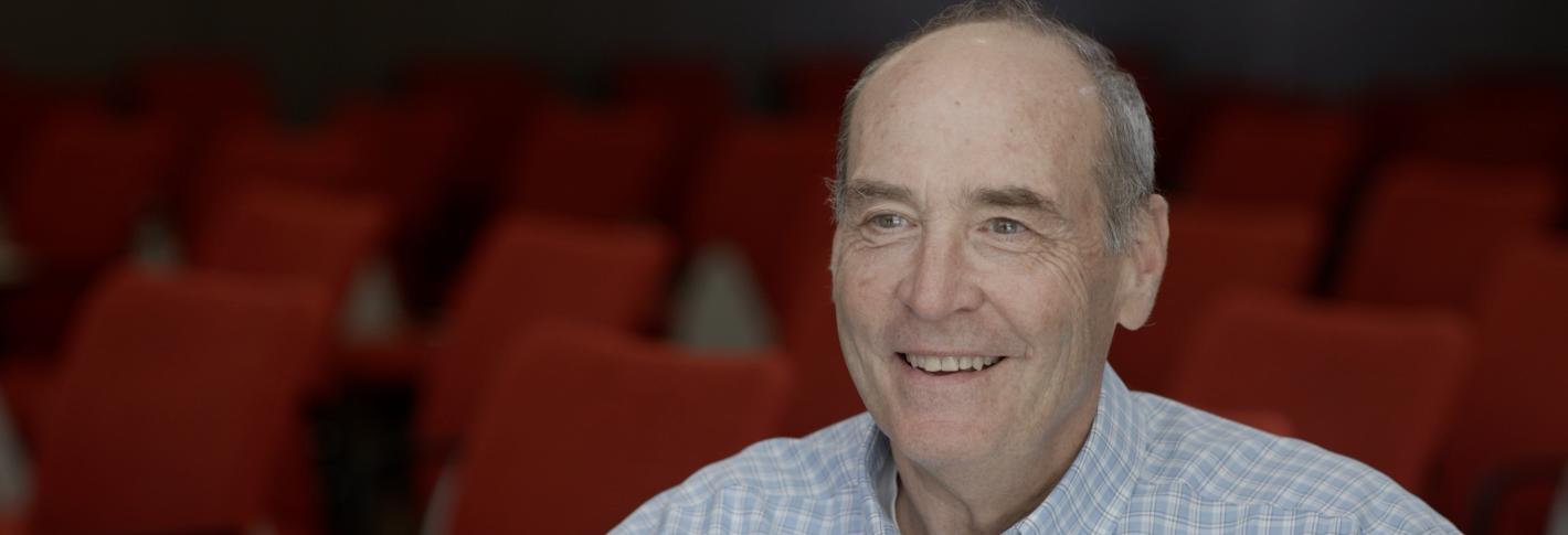 Mark Bear, shown from mid-chest up, smiles as he sits in a lecture room full of red chairs in the backgorund