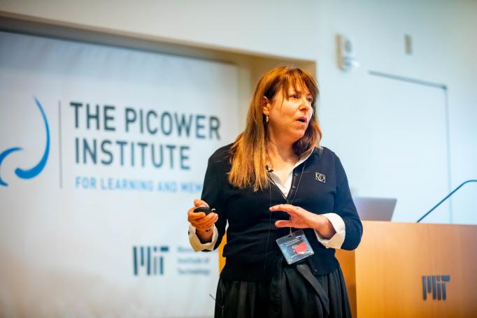 Elly Nedivi stands before the podium and a Picower logo sign as she delivers her talk